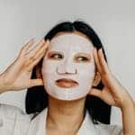 Asian woman applying hydrating sheet mask as part of her skincare routine.