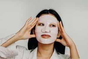 Asian woman applying hydrating sheet mask as part of her skincare routine.